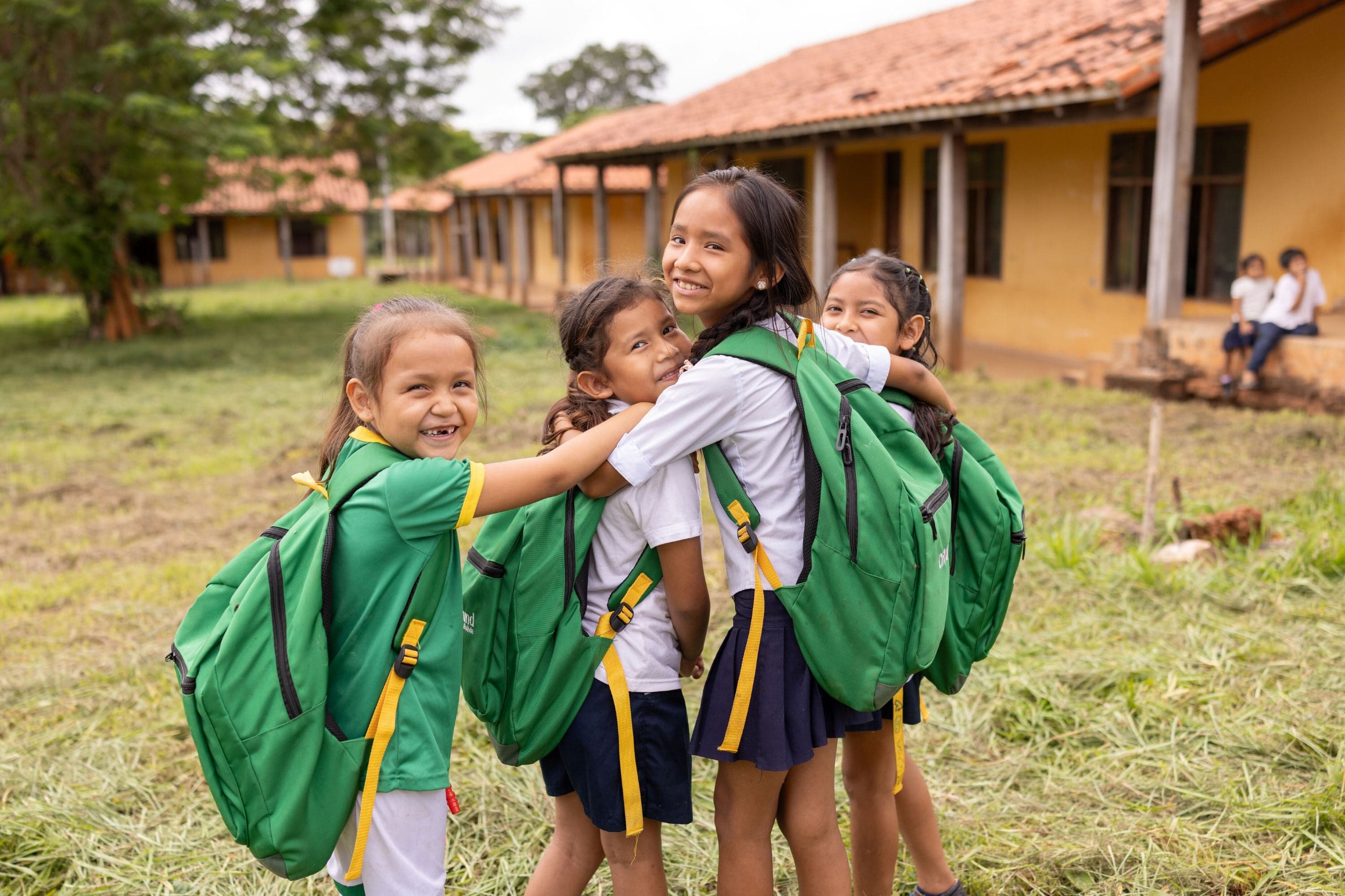 Four schoolgirls in Bolivia wearing green backpacks look back at the camera while placing their arms over each other as they stand at their school.